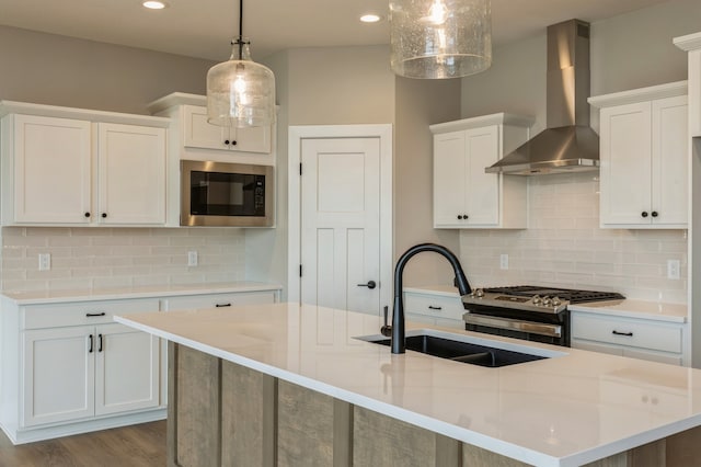 kitchen featuring wall chimney range hood, an island with sink, white cabinetry, stainless steel appliances, and pendant lighting