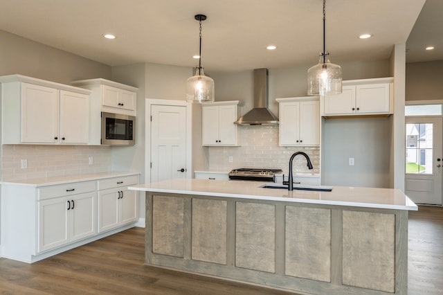 kitchen featuring wall chimney range hood, stainless steel appliances, a center island with sink, pendant lighting, and white cabinetry