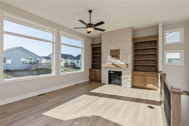 unfurnished living room with a stone fireplace, ceiling fan, built in features, and wood-type flooring