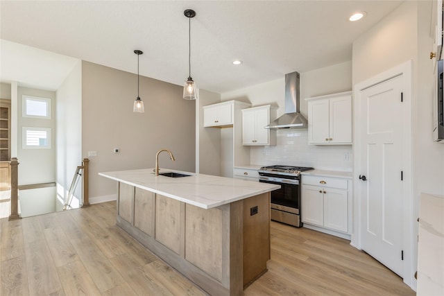 kitchen with wall chimney exhaust hood, gas range, sink, white cabinets, and an island with sink