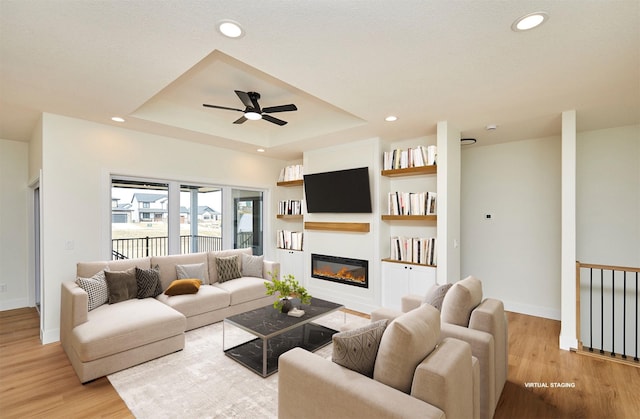 living area featuring a tray ceiling, recessed lighting, a glass covered fireplace, and light wood-style floors