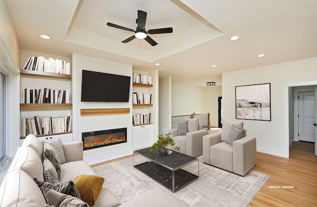 living room featuring built in shelves, recessed lighting, wood finished floors, a raised ceiling, and a glass covered fireplace