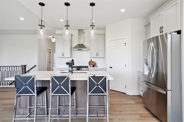 kitchen featuring white cabinetry, stainless steel refrigerator with ice dispenser, a kitchen island with sink, sink, and wall chimney range hood