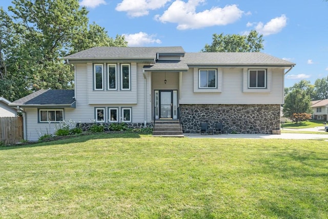 bi-level home featuring stone siding, a front lawn, a shingled roof, and fence