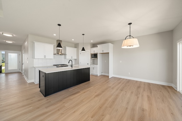 kitchen with stainless steel microwave, wall chimney range hood, a center island with sink, white cabinetry, and hanging light fixtures