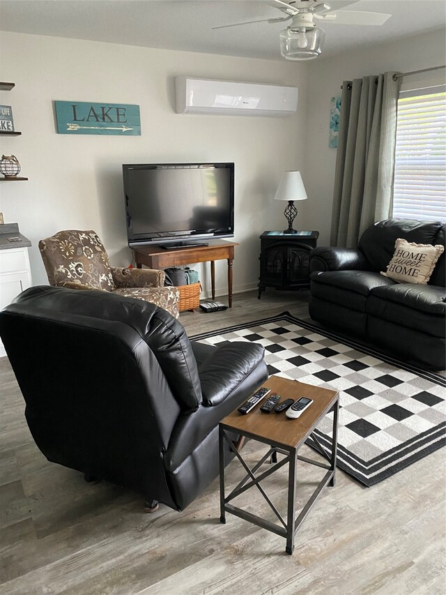 living room with a wall mounted air conditioner, a wood stove, wood finished floors, and a ceiling fan