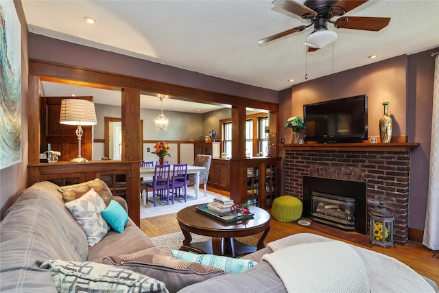 living room with light hardwood / wood-style floors, ceiling fan with notable chandelier, a textured ceiling, and a brick fireplace