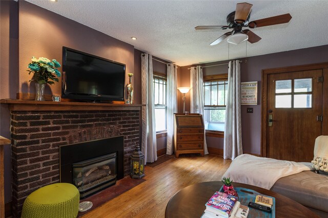 living room featuring ceiling fan, a textured ceiling, a brick fireplace, wood finished floors, and baseboards