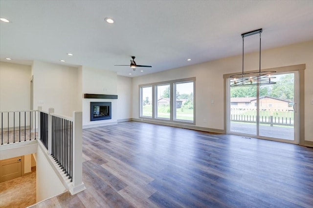 unfurnished living room featuring baseboards, a glass covered fireplace, wood finished floors, ceiling fan with notable chandelier, and recessed lighting