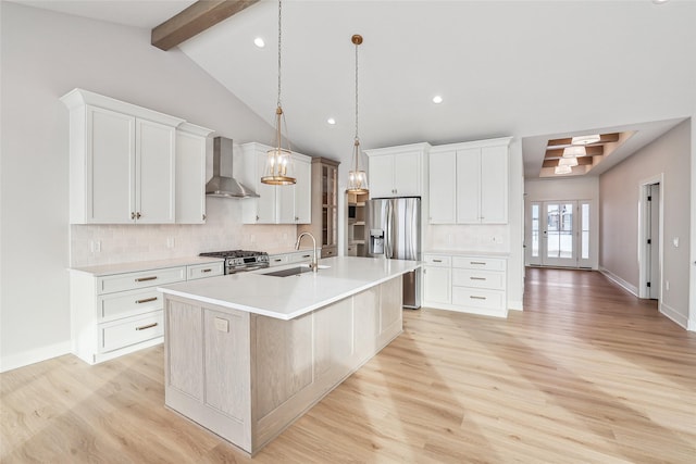 kitchen featuring stainless steel appliances, wall chimney range hood, decorative light fixtures, a kitchen island with sink, and white cabinets