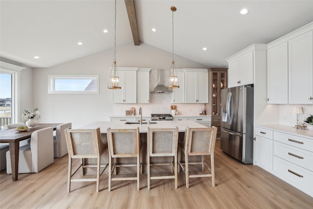 kitchen featuring decorative light fixtures, stainless steel refrigerator with ice dispenser, light countertops, a kitchen island with sink, and wall chimney exhaust hood