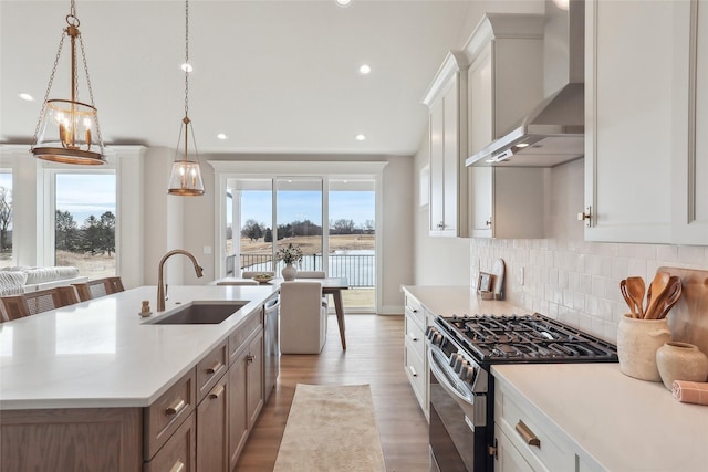 kitchen with a kitchen island with sink, a sink, light countertops, appliances with stainless steel finishes, and wall chimney exhaust hood