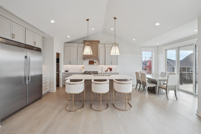 kitchen with an island with sink, light wood-style flooring, stainless steel built in fridge, light countertops, and a sink