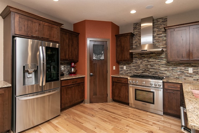 kitchen featuring backsplash, light hardwood / wood-style floors, stainless steel appliances, light stone counters, and wall chimney range hood