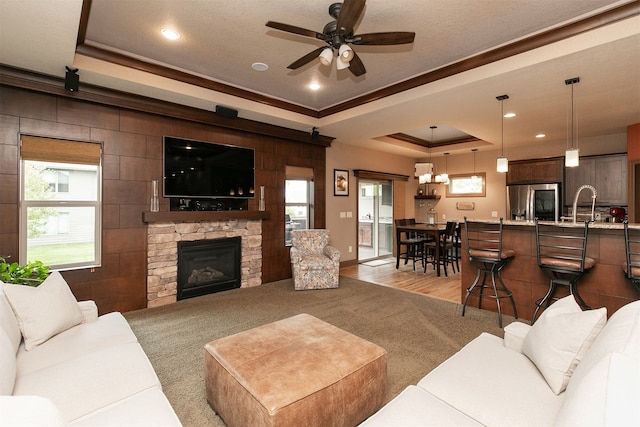 living room with light hardwood / wood-style floors, a tray ceiling, a stone fireplace, ceiling fan, and ornamental molding