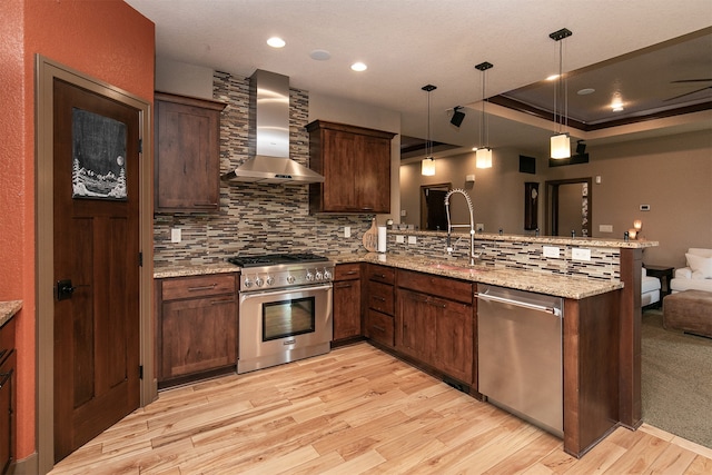 kitchen with light wood-type flooring, pendant lighting, stainless steel appliances, kitchen peninsula, and wall chimney range hood