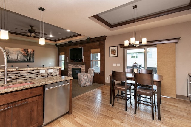 kitchen featuring dishwasher, light hardwood / wood-style flooring, ceiling fan with notable chandelier, a tray ceiling, and a stone fireplace