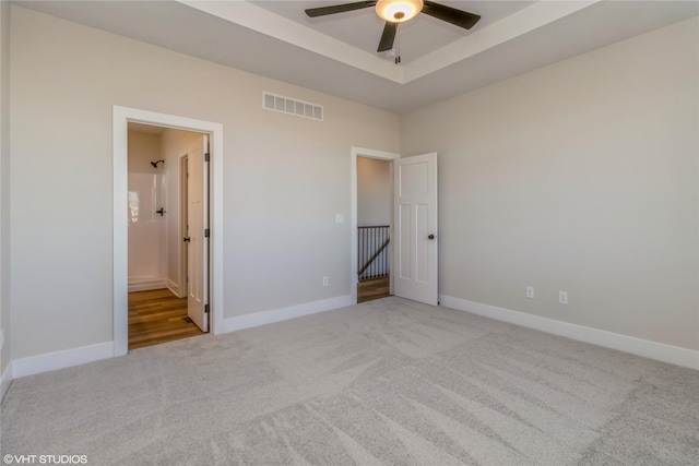 unfurnished bedroom featuring ceiling fan, ensuite bathroom, a tray ceiling, and light carpet