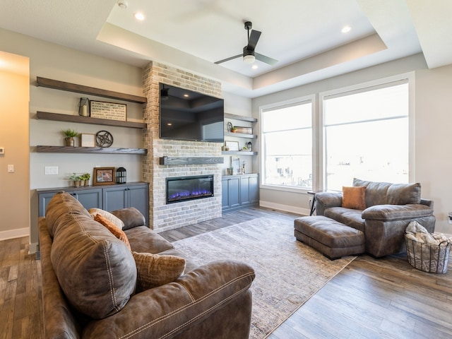 living room featuring a tray ceiling, a brick fireplace, wood-type flooring, and ceiling fan