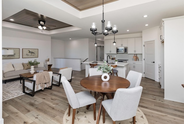 dining area with ceiling fan with notable chandelier, light hardwood / wood-style floors, sink, and a tray ceiling