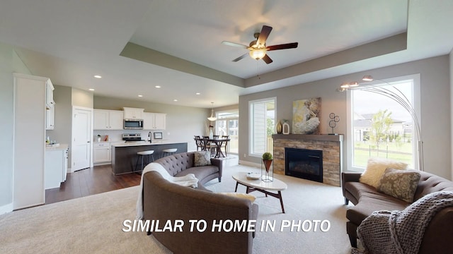 living room with dark wood-type flooring, ceiling fan, a stone fireplace, and a tray ceiling