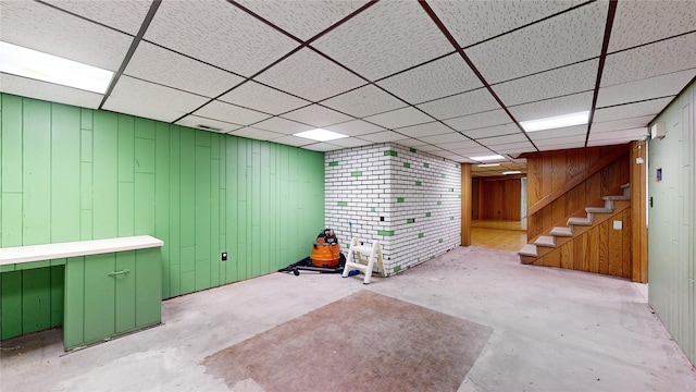 finished basement featuring a paneled ceiling, wooden walls, and stairs