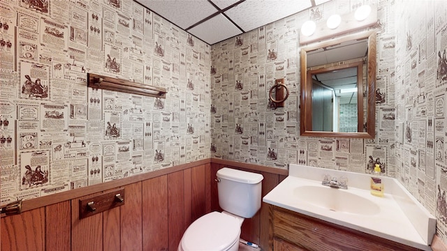 half bath featuring a drop ceiling, toilet, a wainscoted wall, vanity, and wallpapered walls