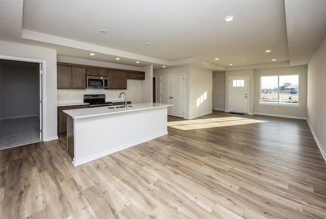 kitchen with dark brown cabinetry, light wood-type flooring, a tray ceiling, an island with sink, and stainless steel appliances