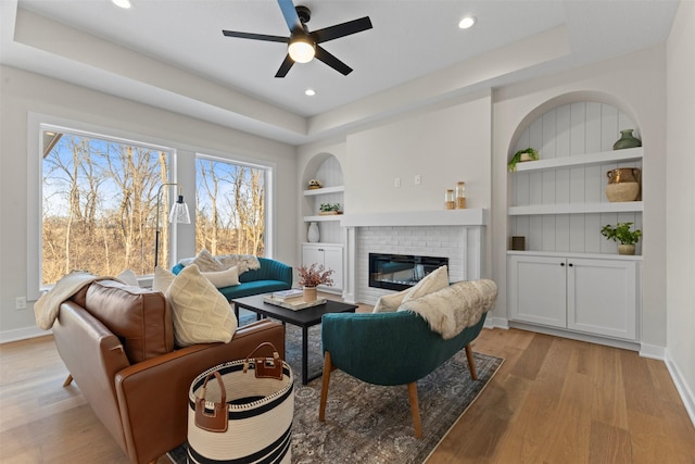 living room featuring ceiling fan, a brick fireplace, a tray ceiling, light hardwood / wood-style flooring, and built in shelves