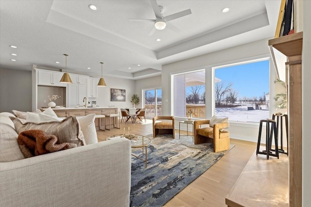 living room with sink, light hardwood / wood-style flooring, ceiling fan, a tray ceiling, and a textured ceiling