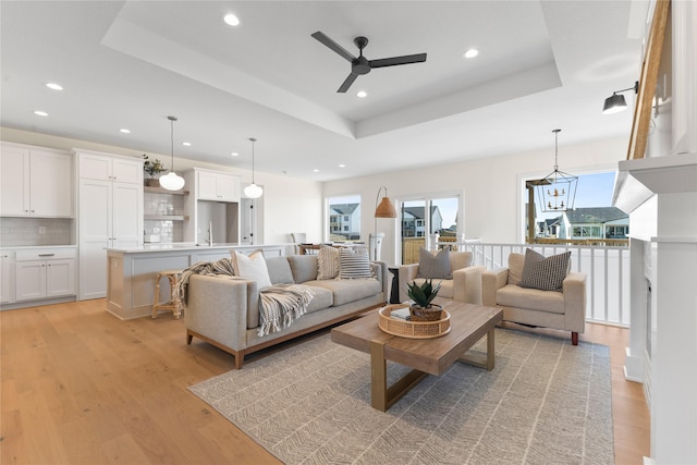 living room with light wood-type flooring, ceiling fan with notable chandelier, and a tray ceiling