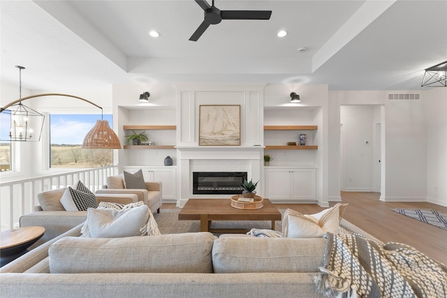 living room featuring ceiling fan with notable chandelier, a large fireplace, a raised ceiling, and light wood-type flooring