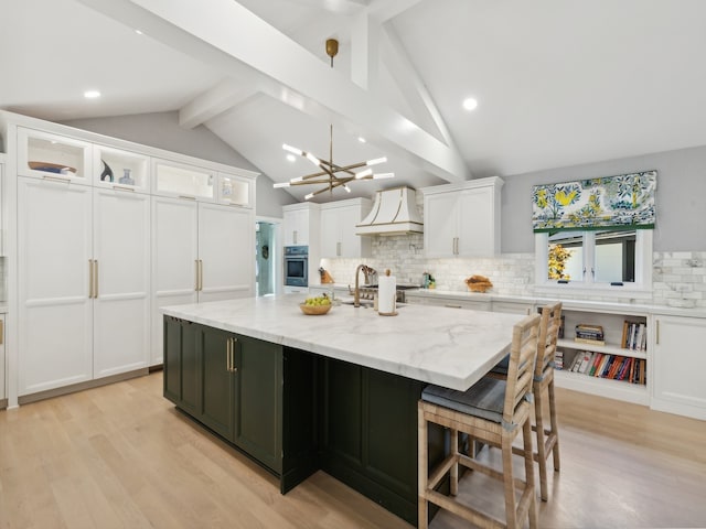 kitchen featuring light wood-type flooring, premium range hood, light stone counters, a center island with sink, and white cabinetry