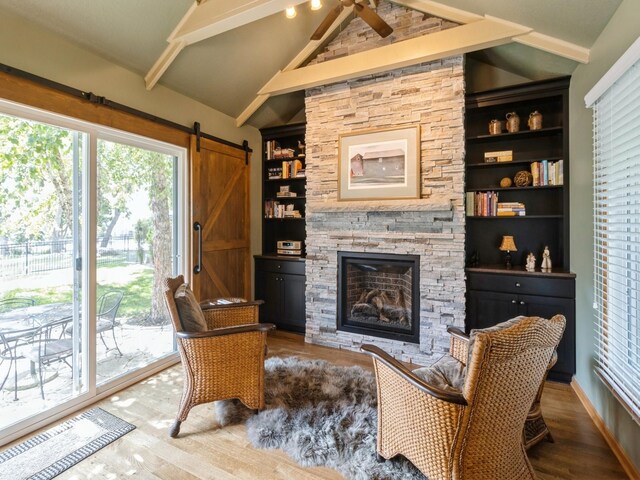 living room with ceiling fan, vaulted ceiling with beams, a stone fireplace, hardwood / wood-style flooring, and a barn door