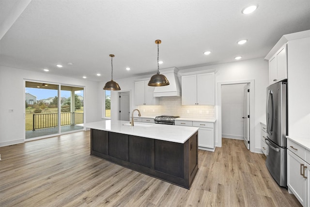 kitchen with appliances with stainless steel finishes, custom exhaust hood, pendant lighting, white cabinetry, and an island with sink