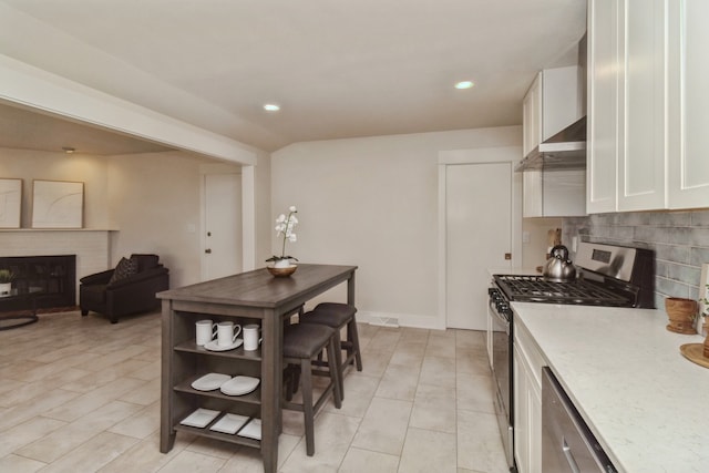 kitchen with a brick fireplace, wall chimney exhaust hood, white cabinetry, stainless steel appliances, and decorative backsplash