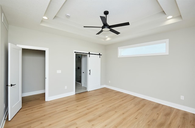 unfurnished bedroom featuring a tray ceiling, a barn door, ceiling fan, and light hardwood / wood-style flooring