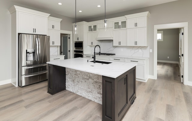 kitchen with light stone countertops, an island with sink, white cabinetry, stainless steel appliances, and custom range hood