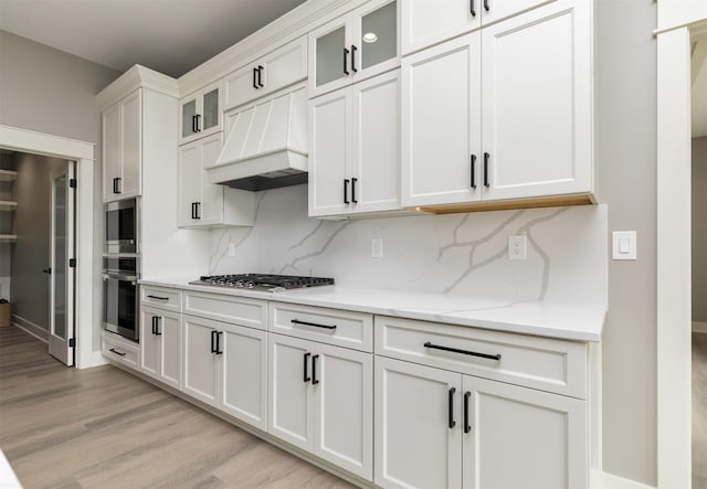 kitchen with tasteful backsplash, light stone countertops, light wood-type flooring, white cabinetry, and custom range hood