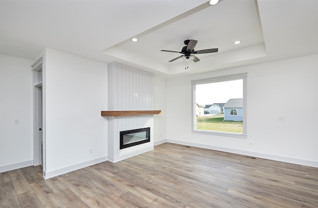 unfurnished living room with a large fireplace, light hardwood / wood-style floors, a raised ceiling, and ceiling fan