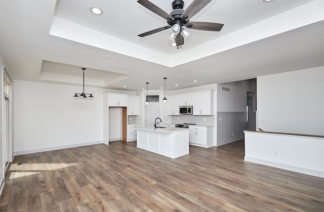 kitchen with a raised ceiling, a center island with sink, stainless steel appliances, hanging light fixtures, and white cabinetry