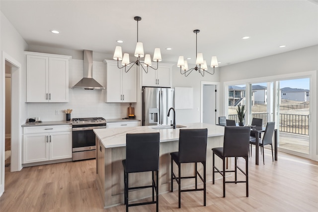 kitchen featuring wall chimney range hood, an island with sink, white cabinets, and appliances with stainless steel finishes