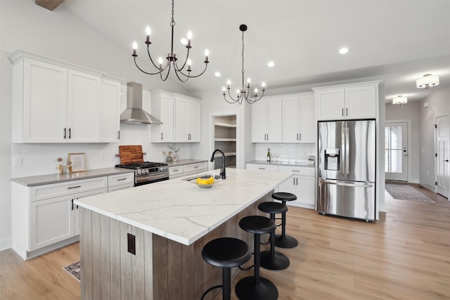 kitchen with wall chimney range hood, sink, an island with sink, white cabinetry, and stainless steel appliances