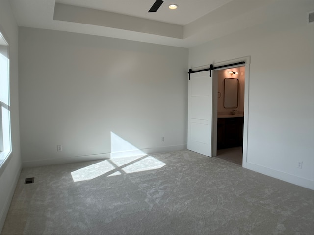 unfurnished bedroom with connected bathroom, a barn door, a raised ceiling, carpet floors, and ceiling fan