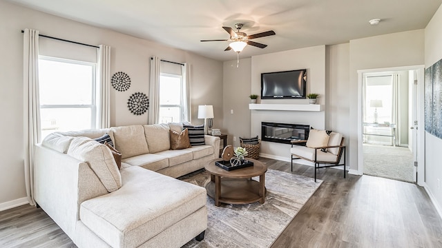 living room featuring a glass covered fireplace, a ceiling fan, baseboards, and wood finished floors