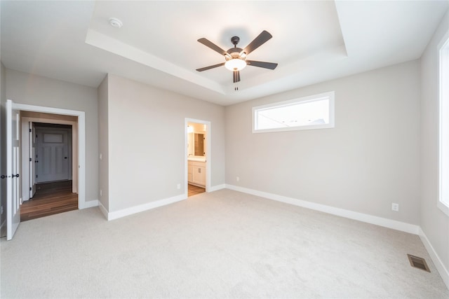 unfurnished bedroom featuring ceiling fan, light colored carpet, a tray ceiling, and ensuite bath