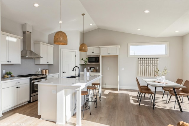 kitchen featuring wall chimney exhaust hood, white cabinetry, appliances with stainless steel finishes, pendant lighting, and a kitchen island with sink