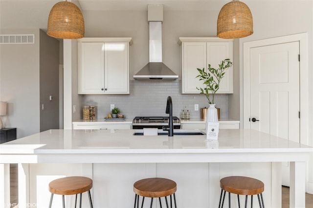 kitchen with a breakfast bar area, white cabinetry, an island with sink, wall chimney range hood, and backsplash