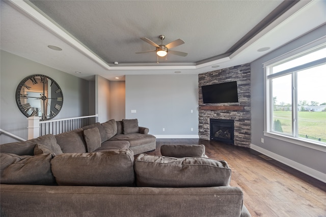 living room with a textured ceiling, light wood-type flooring, a tray ceiling, ceiling fan, and a stone fireplace