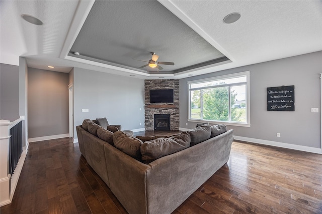 living area featuring a ceiling fan, a stone fireplace, dark wood-style flooring, and a tray ceiling